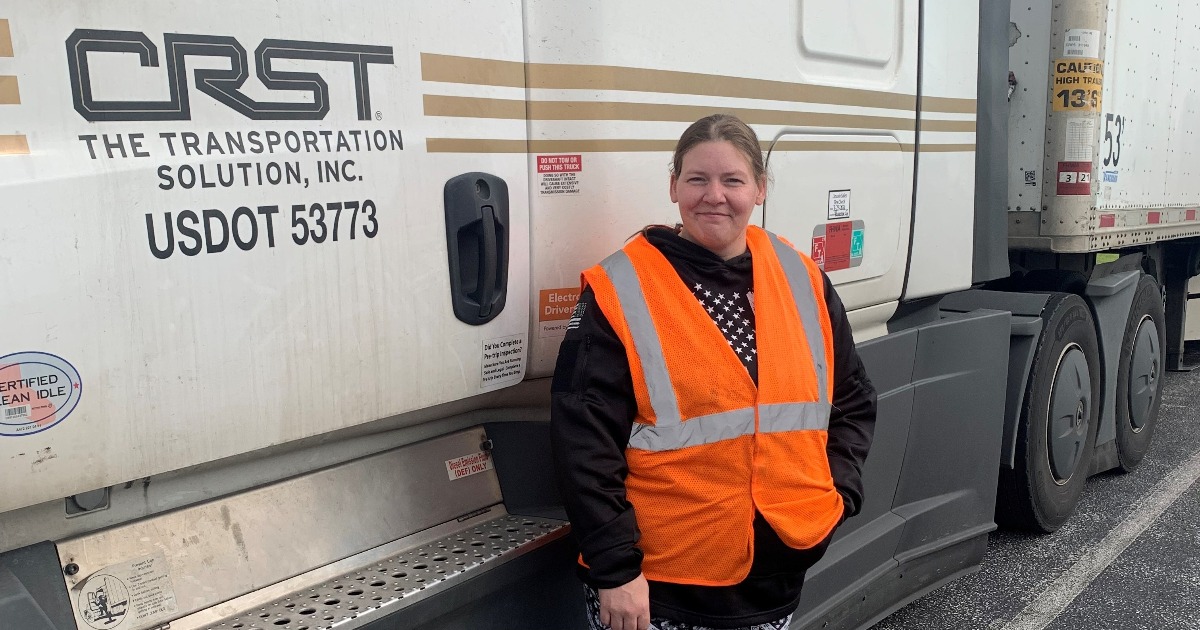 Female CRST driver Shauna Lankford wearing orange safety vest standing on the side of CRST semi truck