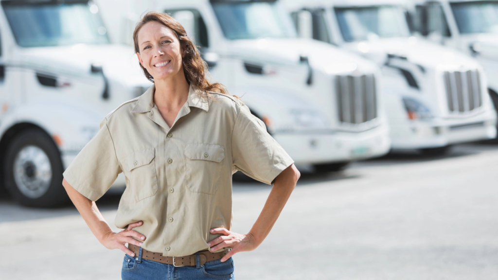 A confident CRST female freight agent stands in front of a row of white semi-trucks, smiling with hands on hips. She represents the logistics professionals who help streamline load booking and optimize freight operations.