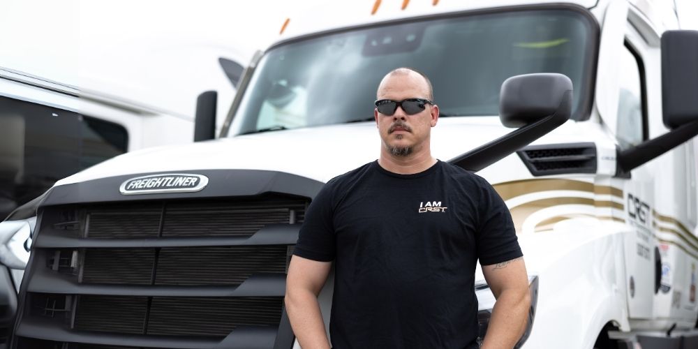 CRST truck driver wearing sunglasses and a black "I AM CRST" t-shirt standing confidently in front of a white Freightliner truck with gold detailing.