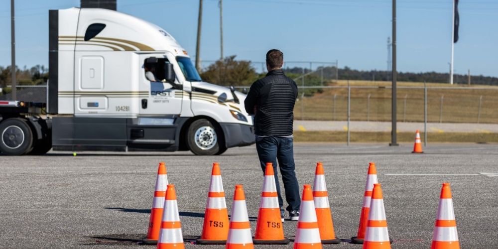 CRST driver training exercise with safety cones in a marked lot. A CRST Freightliner truck waits as an instructor observes the maneuver from behind a formation of bright orange traffic cones.