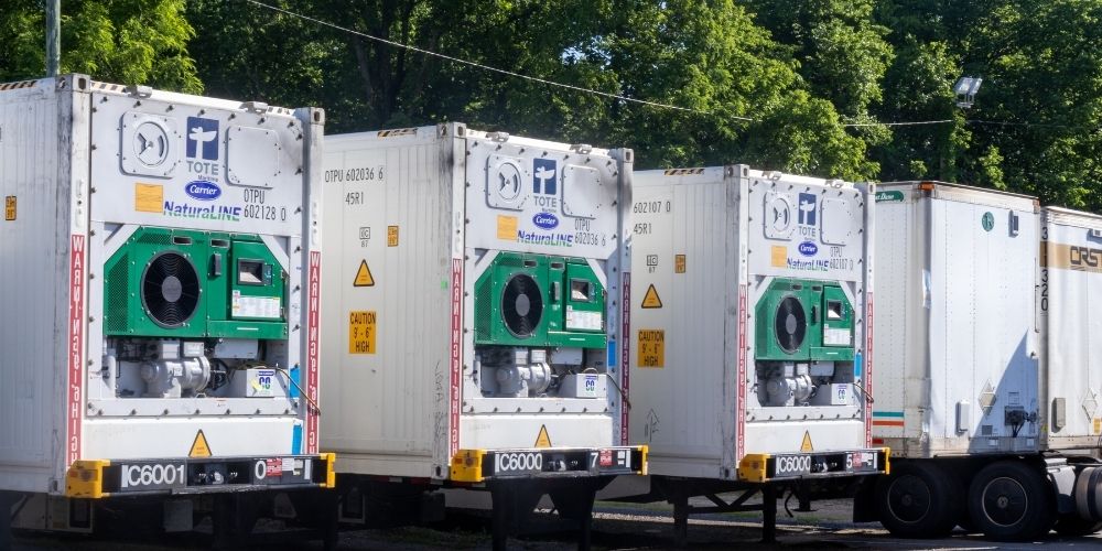 Row of CRST refrigerated shipping containers (reefers) lined up in a lot, including units marked with "Carrier NaturalLINE" and "TOTE," with trees and a light post in the background.