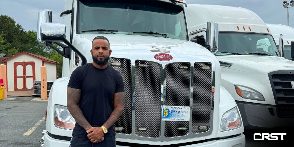 CRST owner-operator Angel Benzant stands confidently in front of his white Peterbilt truck, parked in a lot with other semis