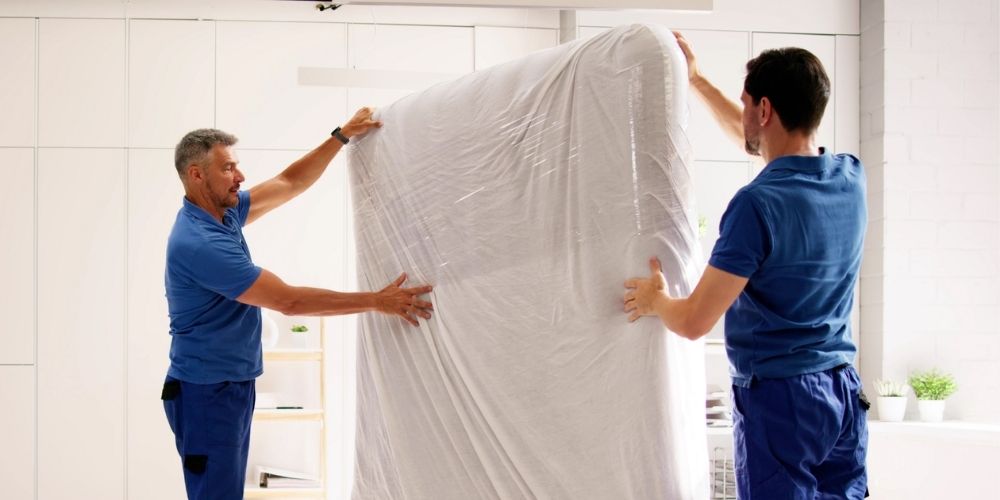 Two movers in blue uniforms carefully lifting and carrying a mattress wrapped in protective plastic inside a modern, well-lit home.