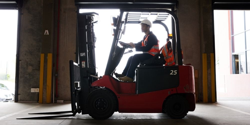 Forklift operator wearing a hard hat and safety vest driving a red forklift inside a warehouse loading area.
