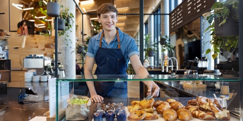 Young male barista wearing a denim apron, smiling while serving pastries at a modern bakery and coffee shop counter, with various baked goods like croissants and muffins displayed in front of him. The café features hanging plants and a stylish, bright interior.