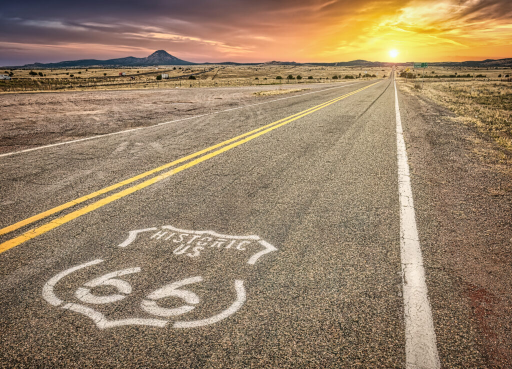 Historic Route 66 shield painted on a two-lane highway stretching toward a sunset over dry Southwestern grassland