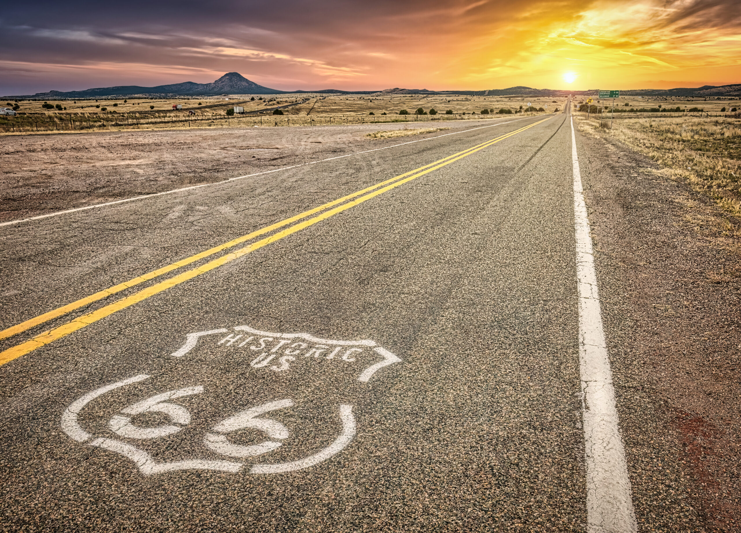Historic Route 66 shield painted on a two-lane highway stretching toward a sunset over dry Southwestern grassland